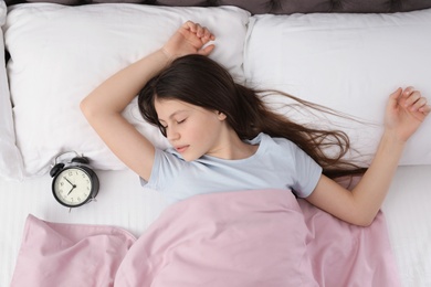 Photo of Beautiful little girl with alarm clock sleeping in bed, top view