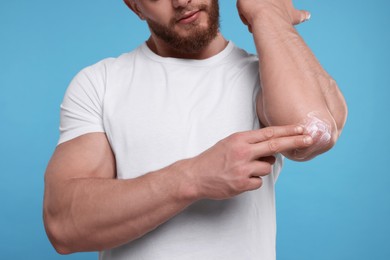 Man applying body cream onto his elbow on light blue background, closeup