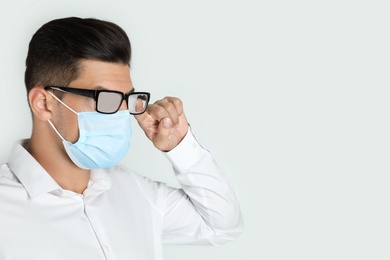 Man wiping foggy glasses caused by wearing medical mask on light background. Space for text