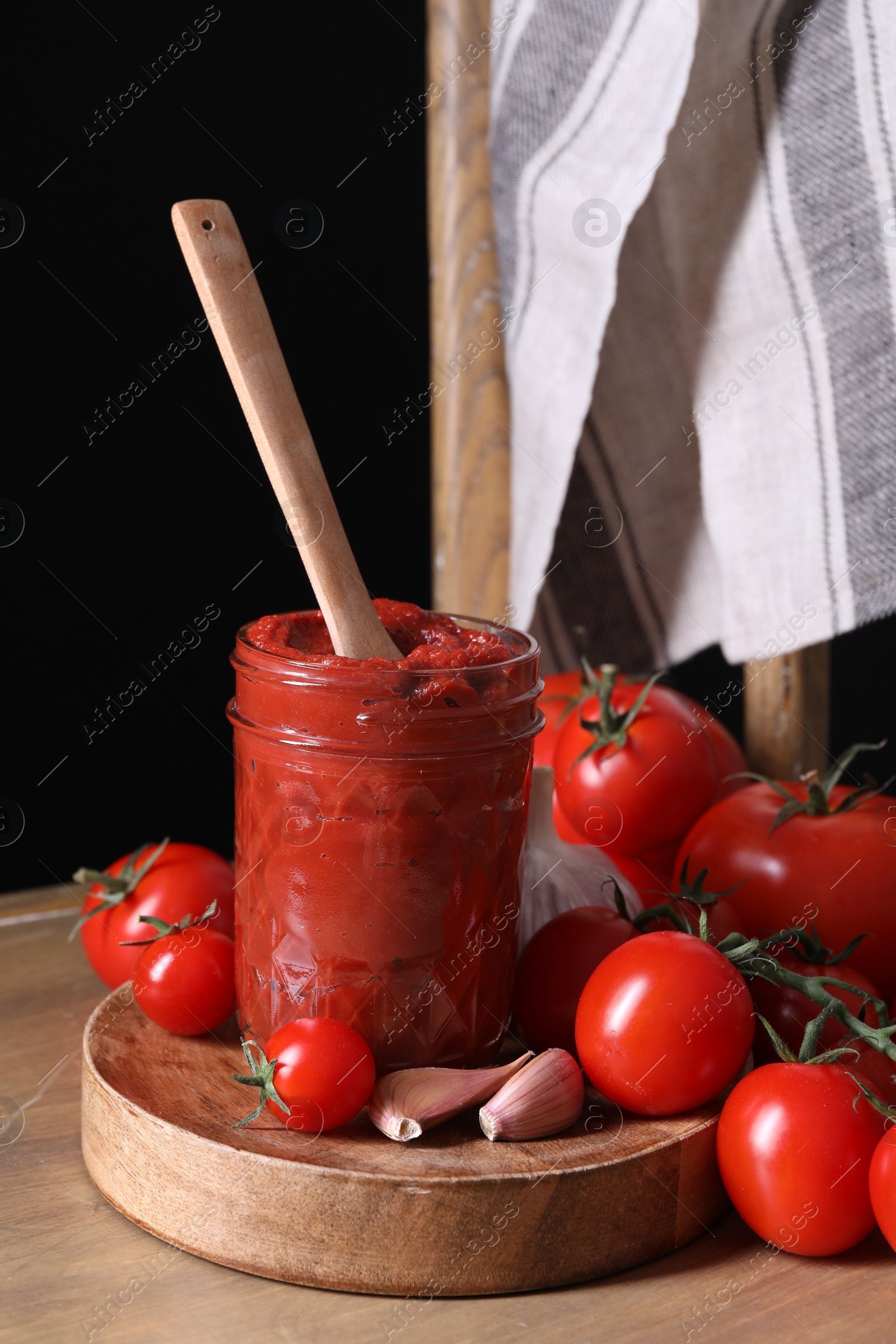 Photo of Jar of tasty tomato paste with spoon and ingredients on wooden table