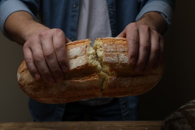 Photo of Man breaking loaf of fresh bread at wooden table, closeup