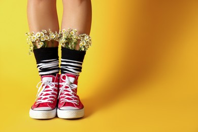 Photo of Woman with beautiful tender chamomile flowers in socks on yellow background, closeup. Space for text