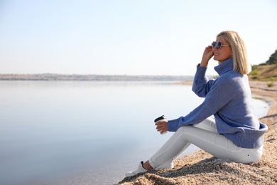 Photo of Happy woman in stylish sweater sitting on beach with cup of coffee
