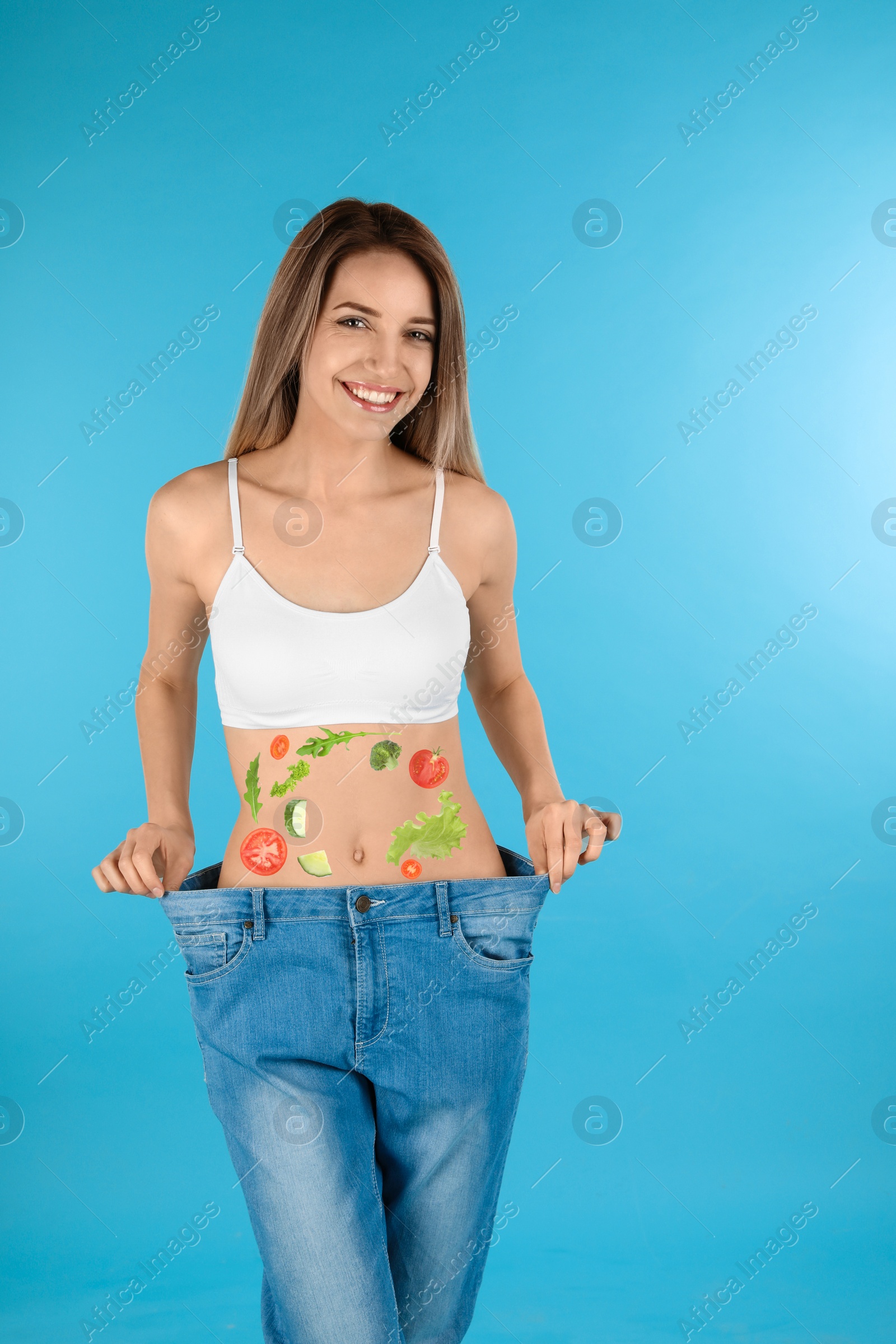 Image of Slim woman in oversized jeans and images of vegetables on her belly against turquoise background. Healthy eating
