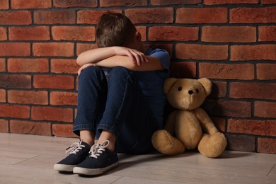 Photo of Child abuse. Upset boy with teddy bear sitting on floor near brick wall indoors