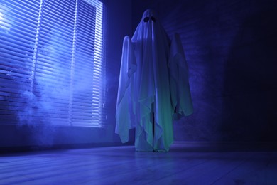 Photo of Creepy ghost. Woman covered with sheet near window in blue light, low angle view