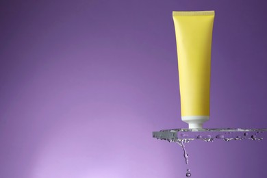 Photo of Moisturizing cream in tube on glass with water drops against violet background. Space for text