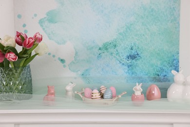 Photo of Easter decorations. Bouquet of tulips in vase, painted eggs and bunny figures on fireplace at home