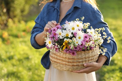 Photo of Woman holding wicker basket with beautiful wild flowers outdoors, closeup