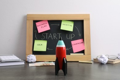 Photo of Blackboard with words Start Up, stationery and toy rocket on grey table