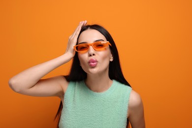 Photo of Beautiful young woman in stylish sunglasses blowing kiss on orange background