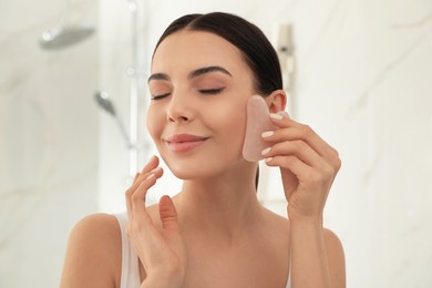 Photo of Beautiful young woman doing facial massage with gua sha tool in bathroom