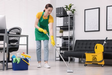 Photo of Cleaning service worker washing floor with mop. Bucket with supplies and wet floor sign in office