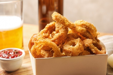 Photo of Cardboard box with crunchy fried onion rings and tomato sauce on wooden board, closeup