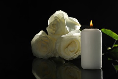 Photo of White roses and burning candle on black mirror surface in darkness, space for text. Funeral symbols