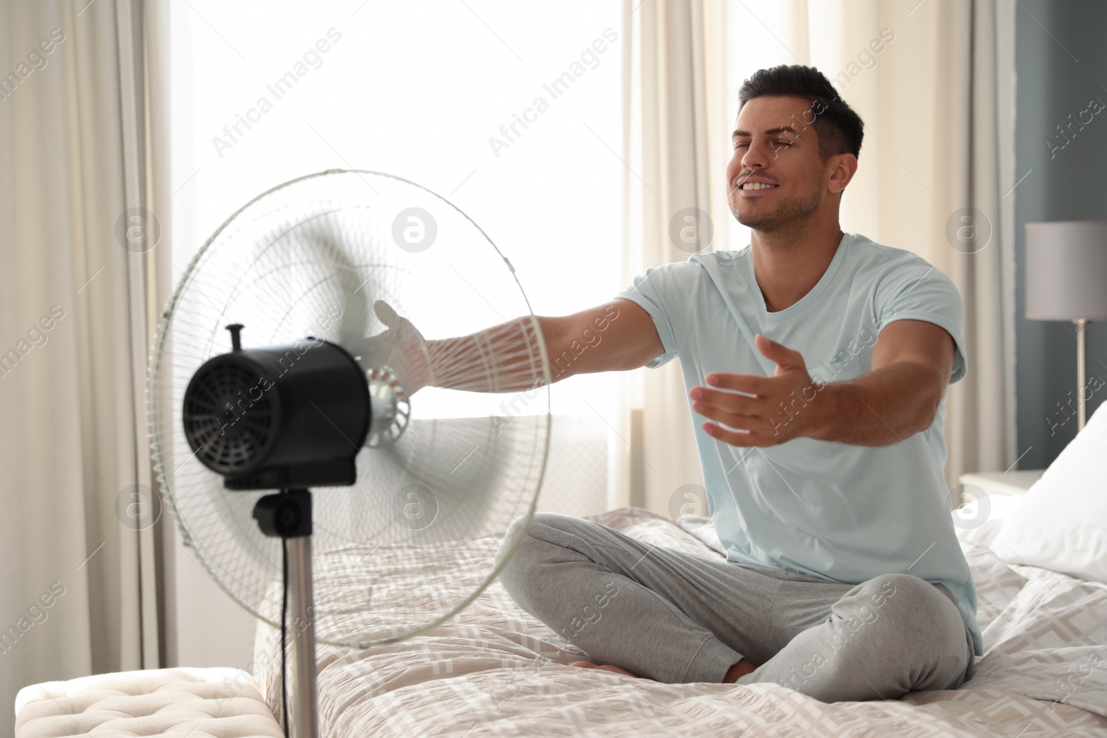 Photo of Man enjoying air flow from fan on bed in room. Summer heat