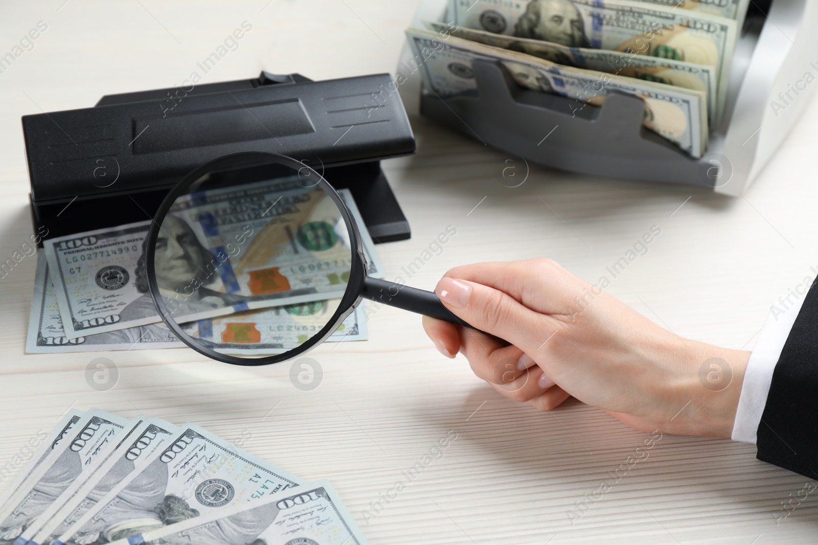 Photo of Woman checking dollar banknotes with currency detector and magnifying glass at white wooden table, closeup. Money examination device