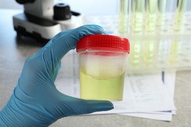 Photo of Doctor holding container with urine sample for analysis at grey table in laboratory, closeup