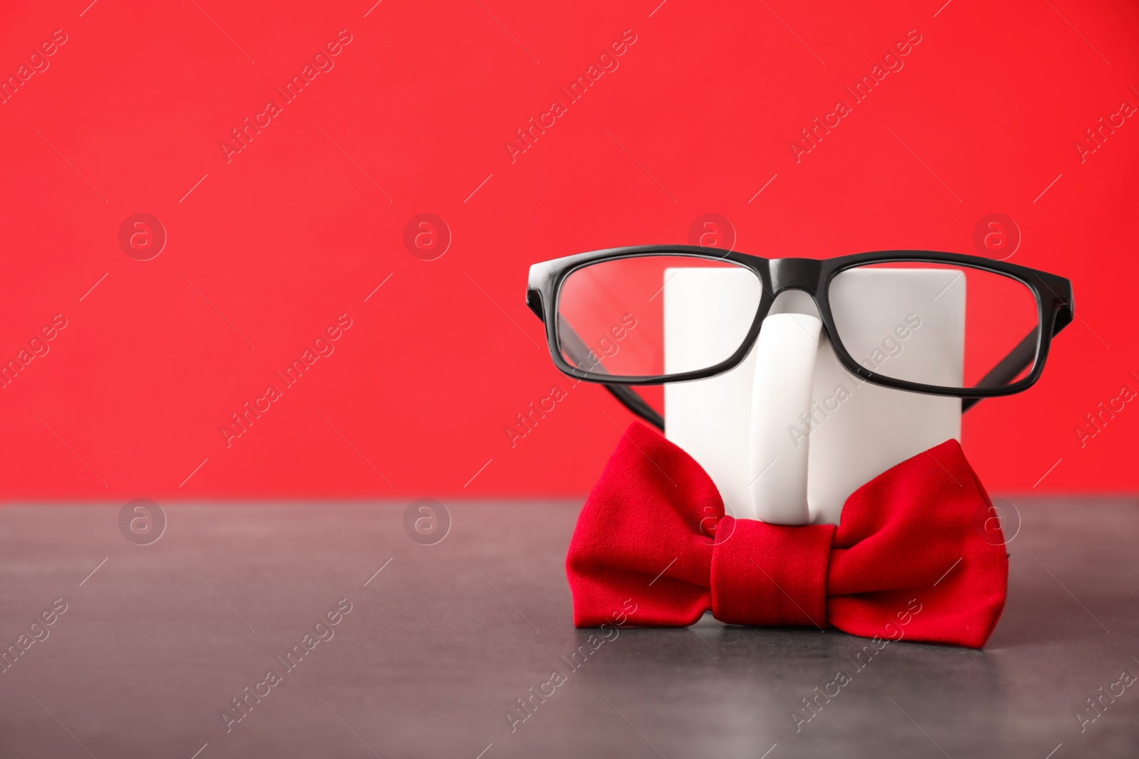 Photo of Funny composition with bow tie, glasses and cup on grey table against red background. Space for text