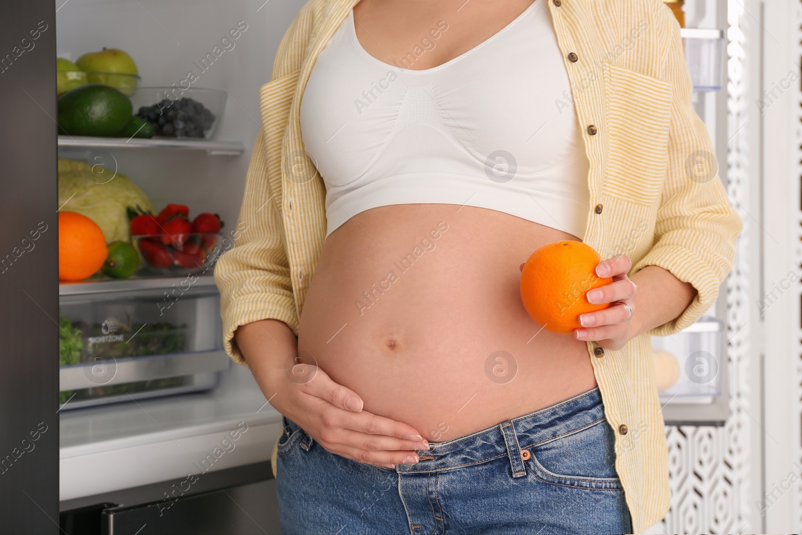 Photo of Young pregnant woman with orange near fridge at home, closeup. Healthy eating