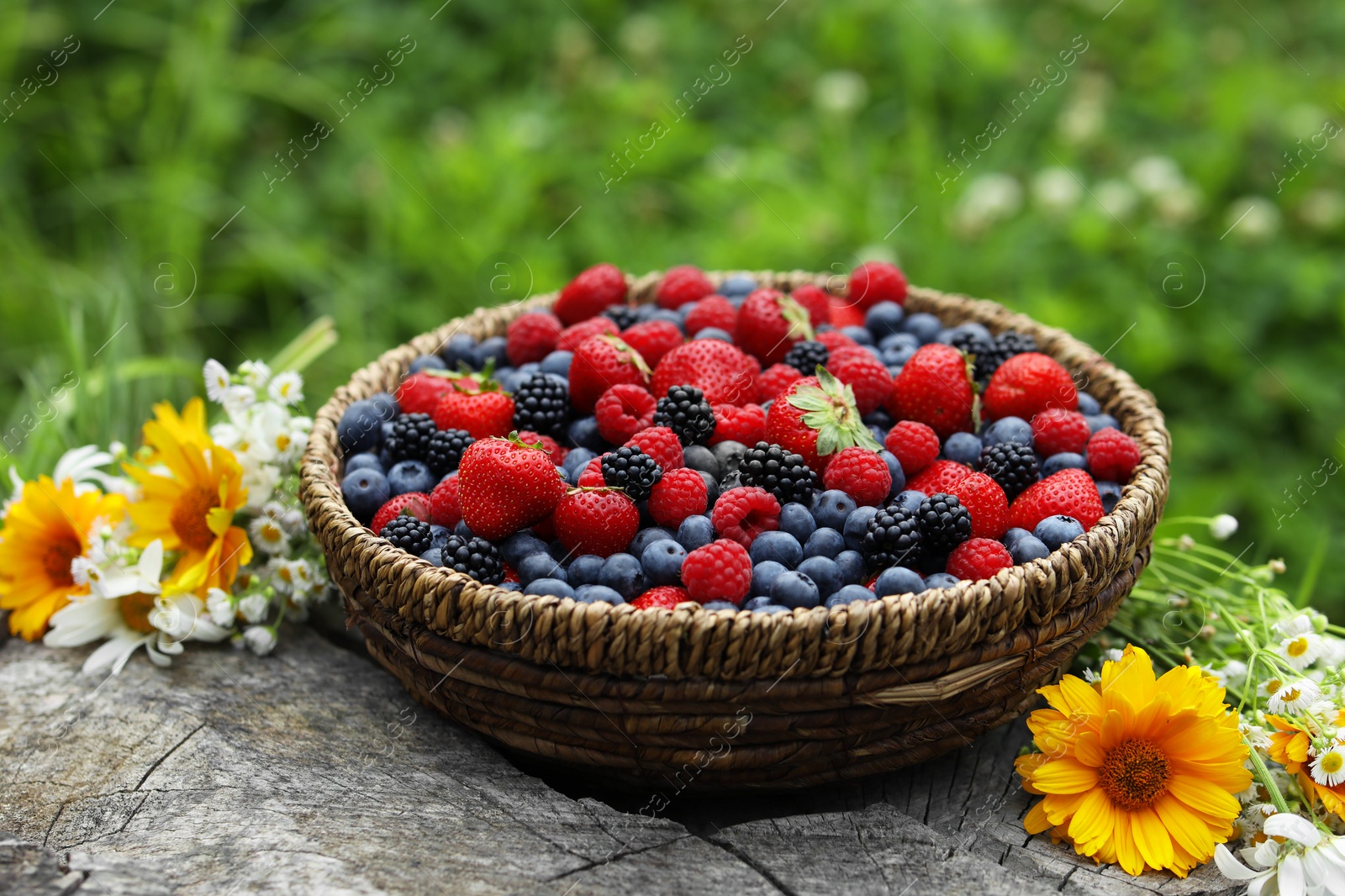 Photo of Wicker bowl with different fresh ripe berries and beautiful flowers on wooden surface outdoors