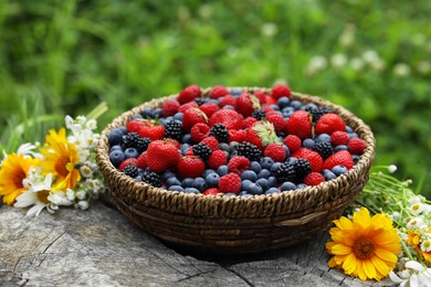 Wicker bowl with different fresh ripe berries and beautiful flowers on wooden surface outdoors