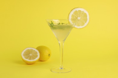 Photo of Martini glass of refreshing cocktail with lemon slice, rosemary and fruits on yellow background