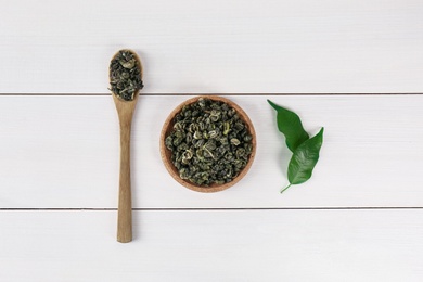 Photo of Dried green tea leaves in spoon and bowl on white wooden table, flat lay