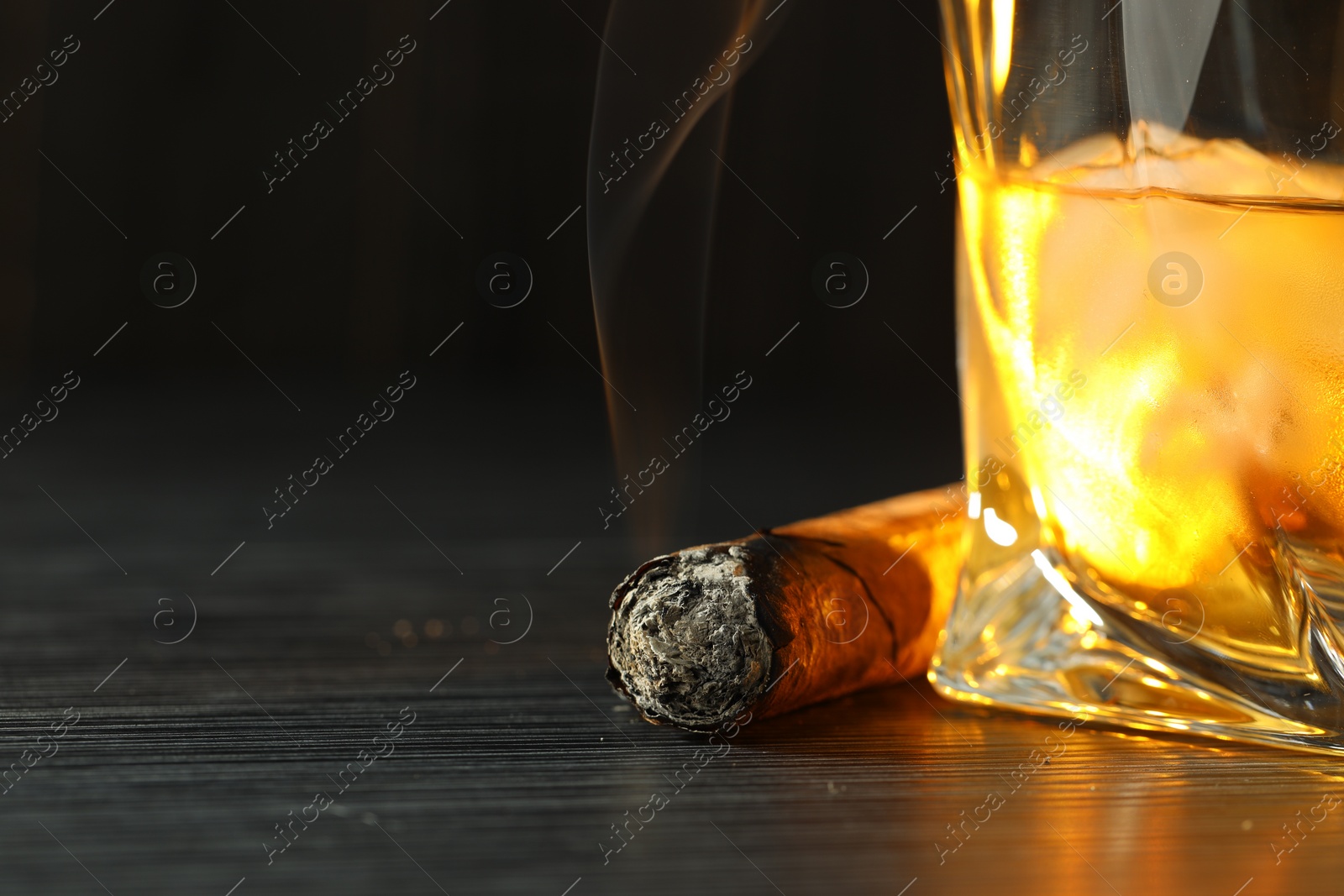 Photo of Glass of whiskey with ice cubes and smoldering cigar on black wooden table, closeup. Space for text