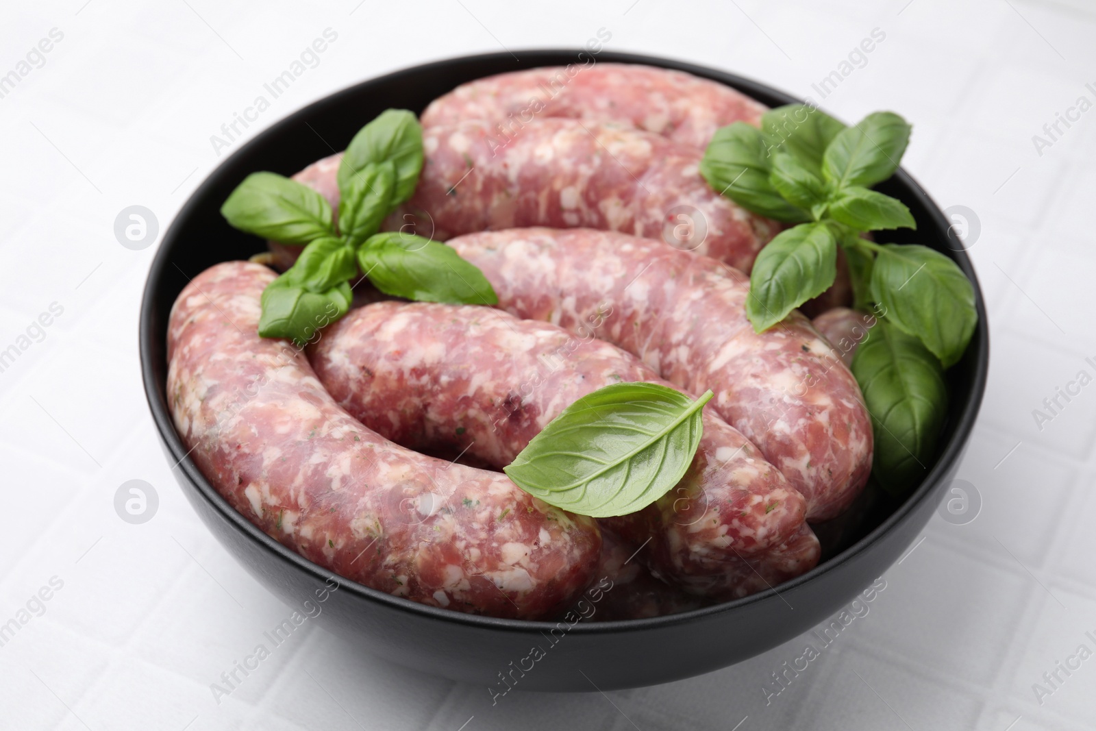 Photo of Raw homemade sausages and basil leaves in bowl on white tiled table, closeup