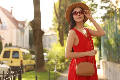 Photo of Beautiful young woman with stylish bag in red dress and sunglasses outdoors, space for text