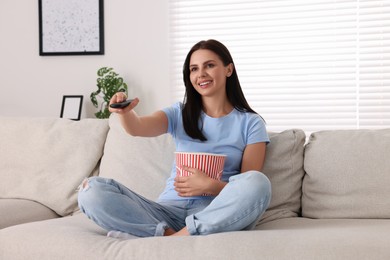 Photo of Happy woman with popcorn bucket changing TV channels with remote control on sofa at home