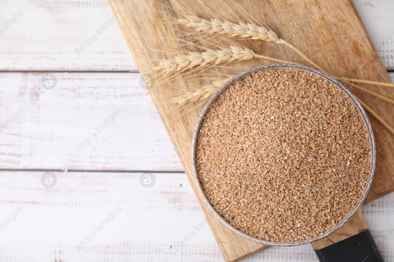 Photo of Dry wheat groats in bowl and spikelets on white wooden table, top view. Space for text