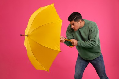 Emotional man with umbrella caught in gust of wind on pink background