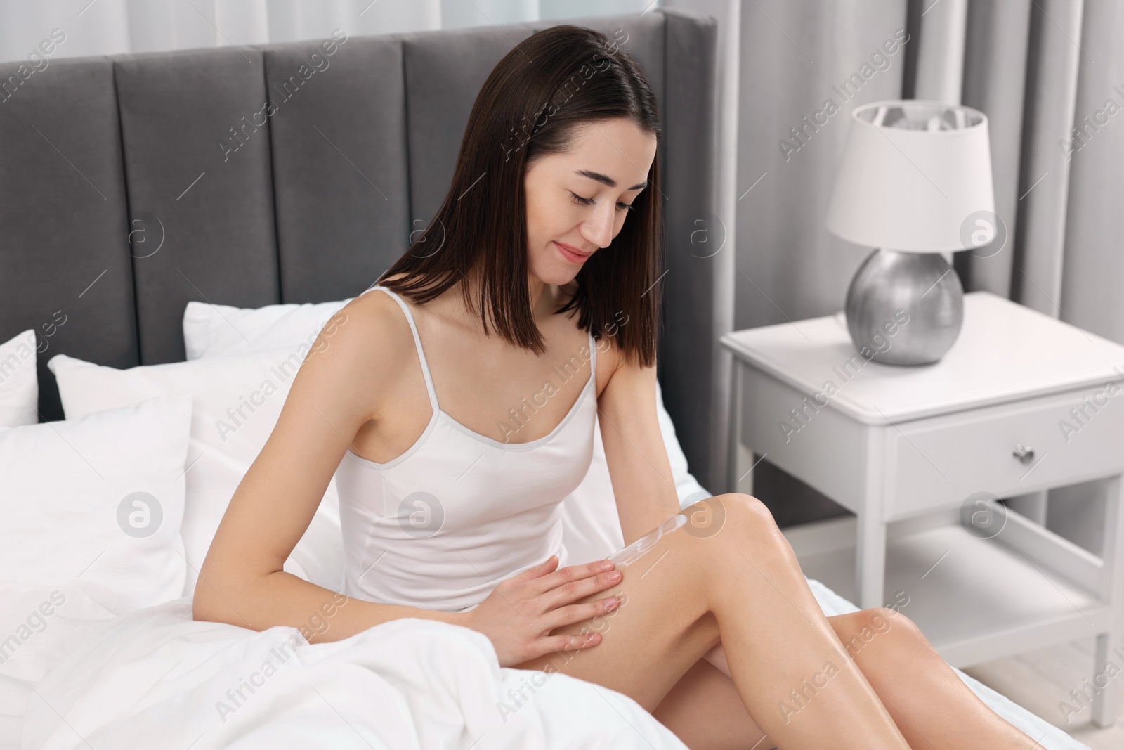 Photo of Young woman applying cream onto leg in bedroom