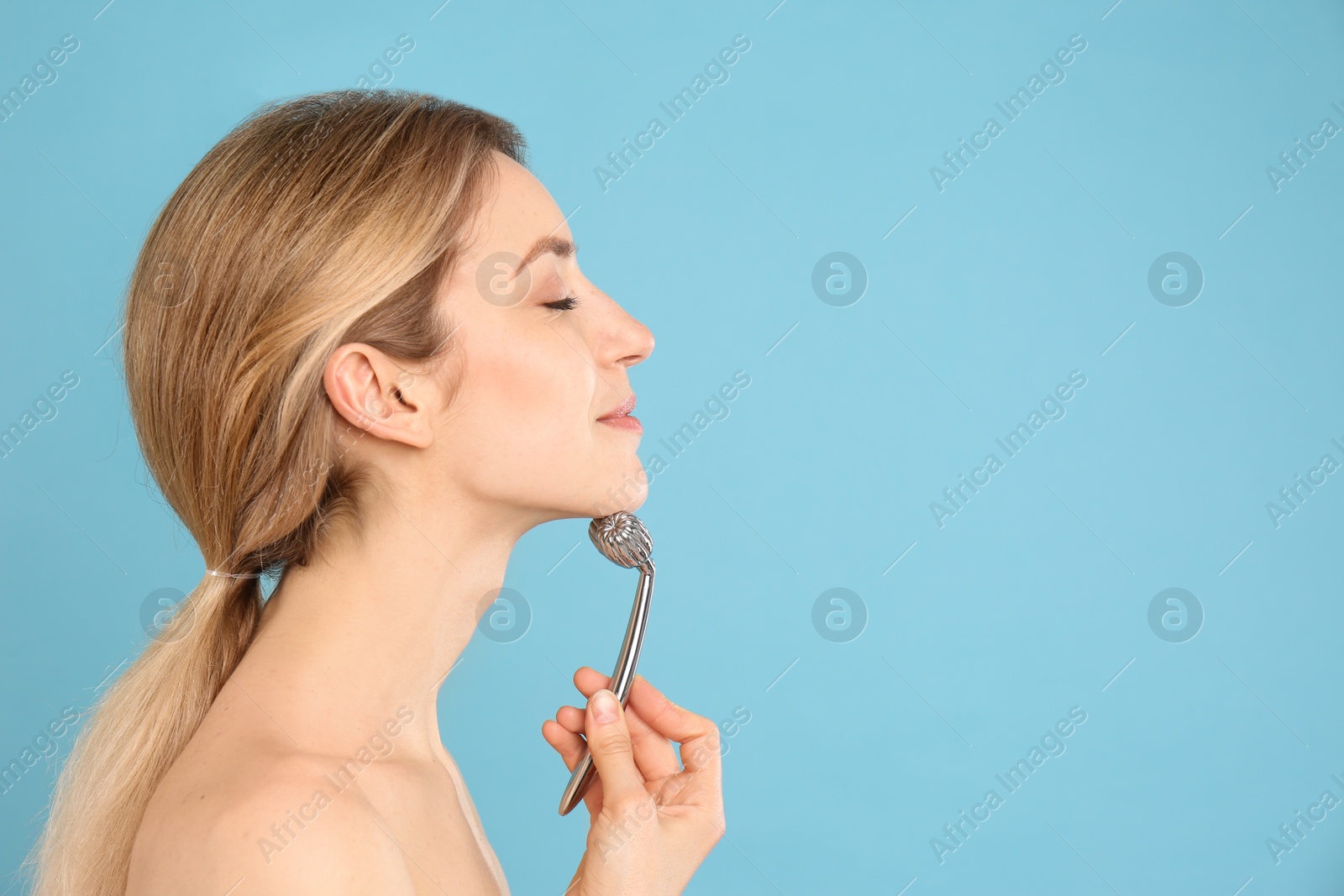 Photo of Young woman using metal face roller on light blue background, space for text