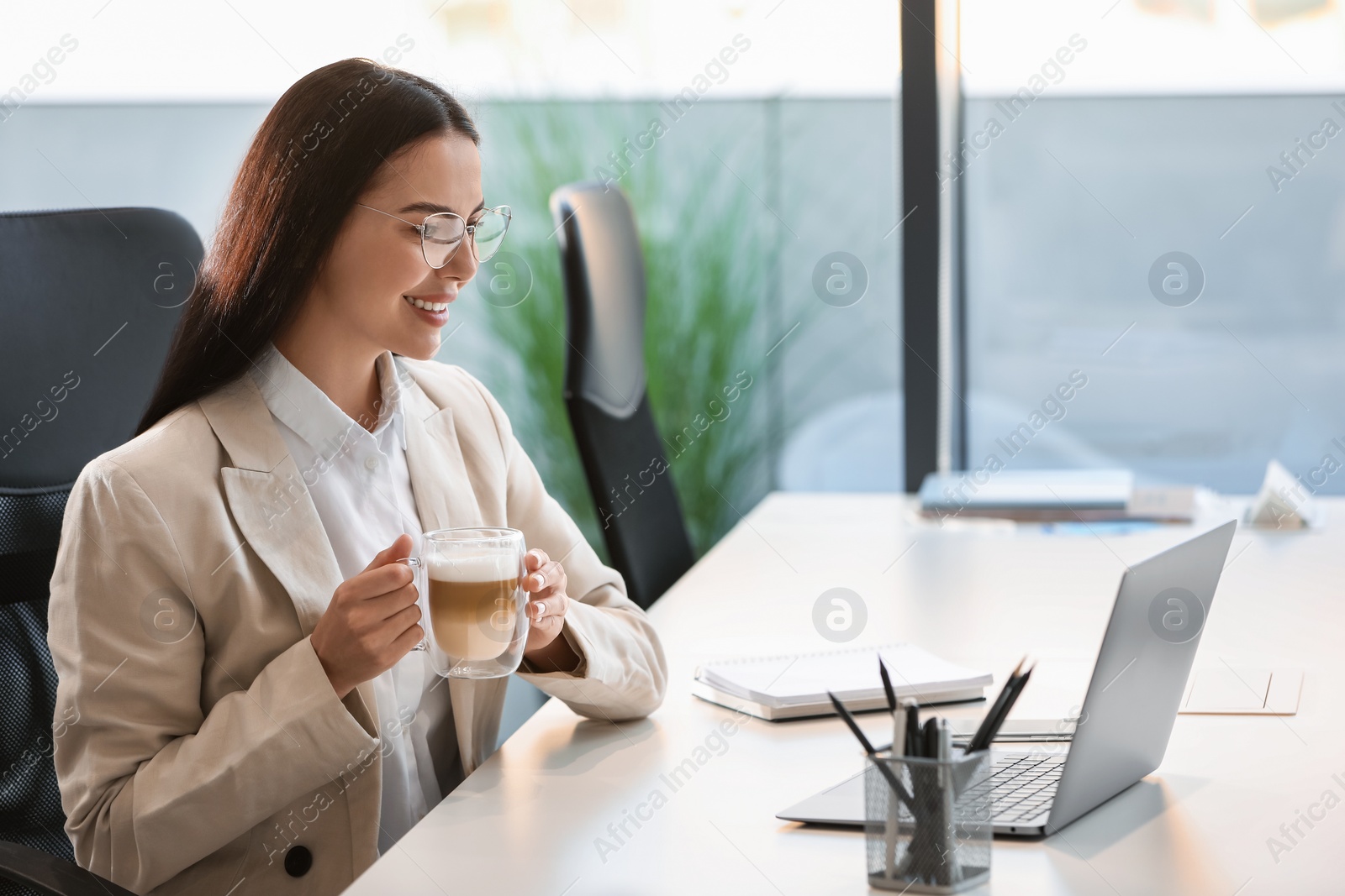 Photo of Happy woman with cup of coffee using modern laptop at white desk in office