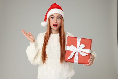 Photo of Emotional young woman in Santa hat with Christmas gift on light grey background