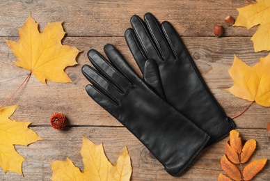 Stylish black leather gloves and dry leaves on wooden table, flat lay