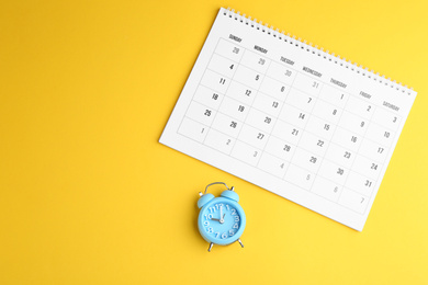 Calendar and alarm clock on yellow background, flat lay. Space for text