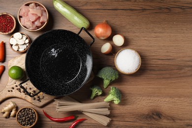 Empty iron wok surrounded by raw ingredients on wooden table, flat lay. Space for text