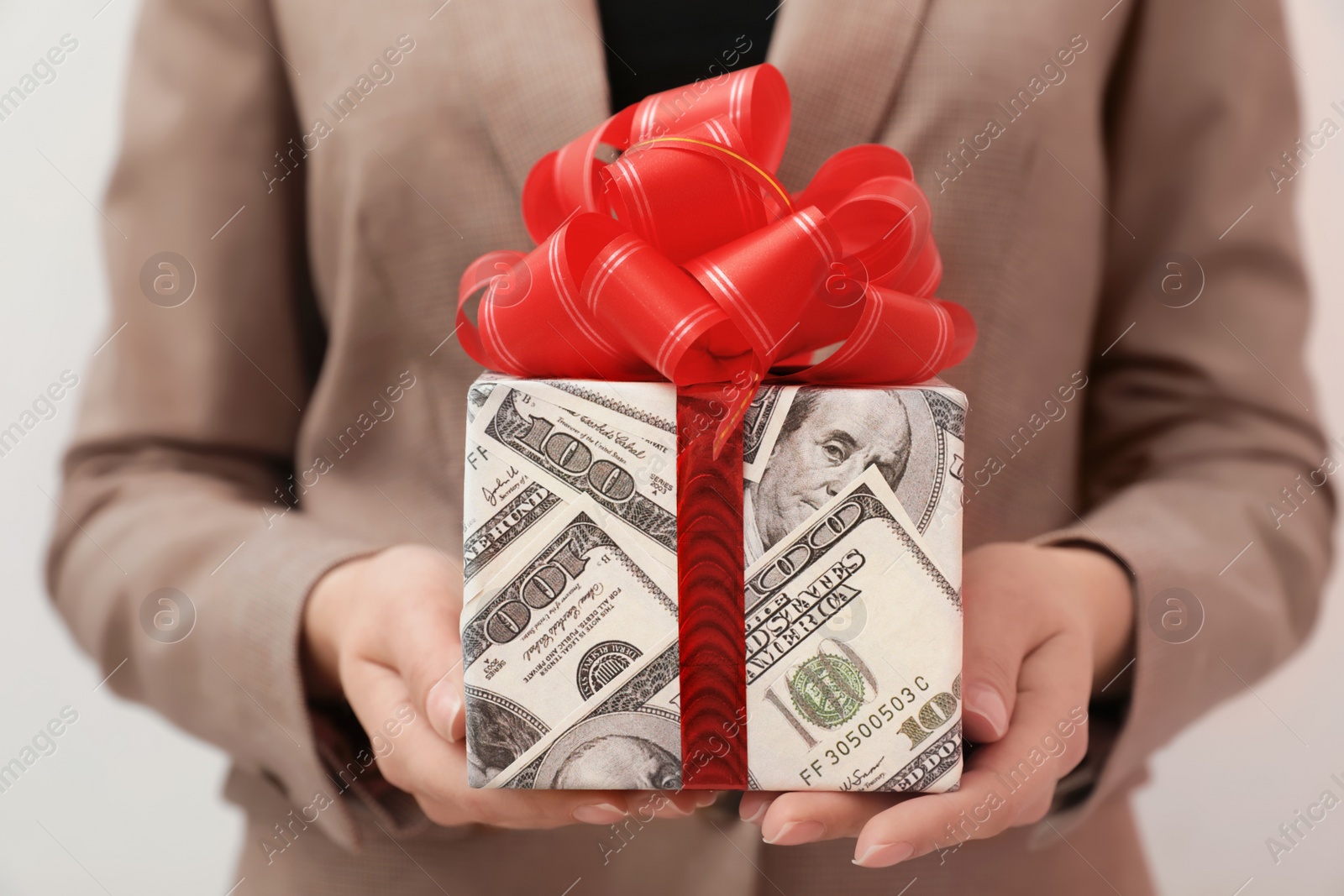 Photo of Woman holding gift box wrapped in dollars on light background, closeup