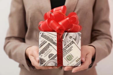 Woman holding gift box wrapped in dollars on light background, closeup