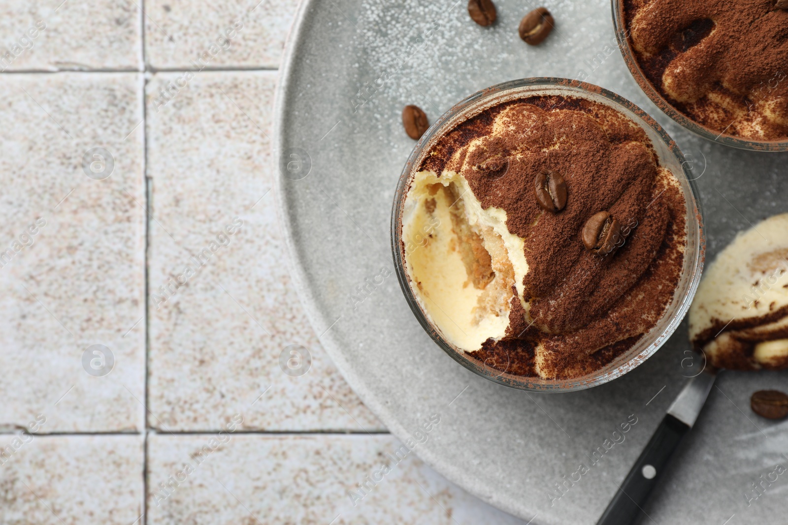 Photo of Delicious tiramisu in glasses, spoon and coffee beans on light tiled table, top view. Space for text