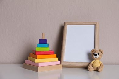 Photo of Empty photo frame, toy bear and pyramid on white table near grey wall. Space for design