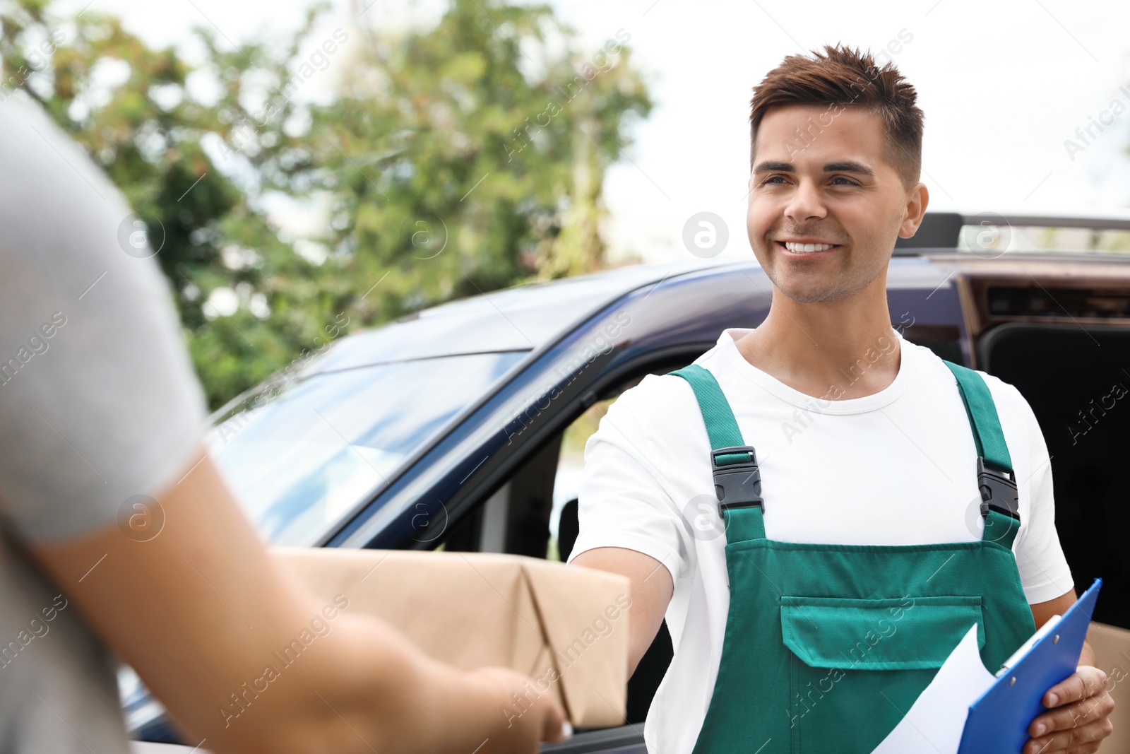 Photo of Young delivery courier with clipboard giving parcel to customer outdoors