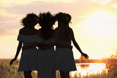 Young friends in wreaths of flowers standing near river, back view. Silhouettes of women at sunrise