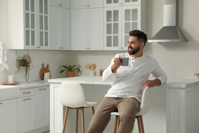 Photo of Handsome young man with cup of tea sitting on stool in kitchen