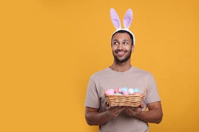 Photo of Happy African American man in bunny ears headband holding wicker tray with Easter eggs on orange background, space for text