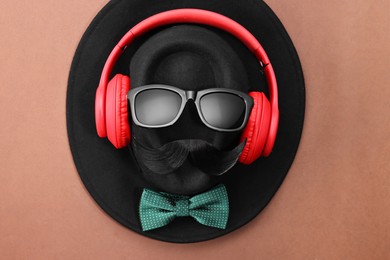 Man's face made of artificial mustache, sunglasses and hat on brown background, top view